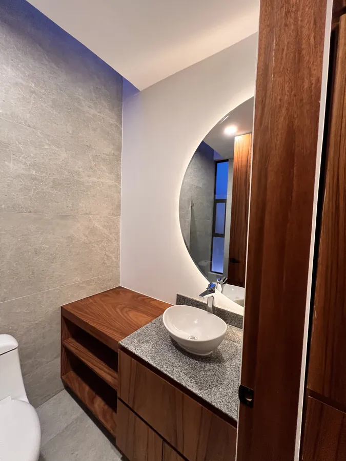 Baño con mueble de madera, lavabo de piedra y espejo circular — residencia contemporánea de doble altura en Puebla