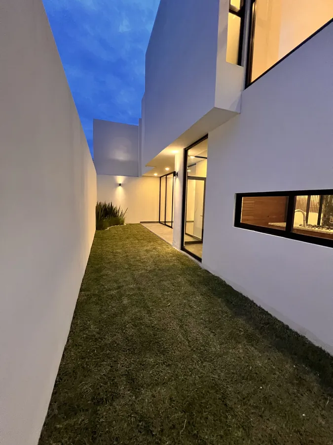 Patio lateral con jardín y fachada minimalista al anochecer — residencia contemporánea de doble altura en Puebla